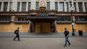 兩名戴著口罩的人走過一間用木板封住的Macy's商店，上方有時鐘。