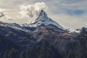 在多雲的天空下，積雪的山峰從森林覆蓋的山坡上拔地而起。