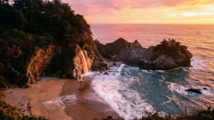 日落時分，瀑布流入海洋，岩石海灘上波浪拍打，懸崖上樹木覆蓋。