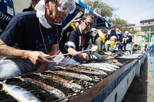 男士們在陽光明媚的戶外節日烤魚，有觀眾觀看。 — 精細 壁紙