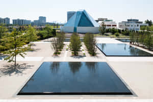現代玻璃金字塔建築，在城市環境中配有倒影池和樹木。 — MacBook 壁紙