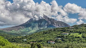 火山在藍天白雲下，於綠色山丘上冒煙，山丘上散落著房屋。