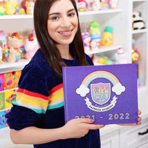 女子手持Rainbow Academy 2021-2022年刊，被色彩繽紛的毛絨玩具環繞。