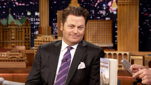 Nick Offerman 身穿西裝，在身後城市燈光的脫口秀舞台上手持自己的書。 — 曉格蘭特 GIF 壁紙