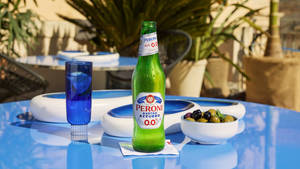 Peroni Nastro Azzurro 0.0 啤酒瓶、玻璃杯和橄欖在戶外藍色桌子上。
