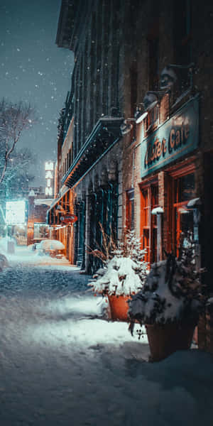 夜間積雪的都市街道，閃爍的咖啡館燈光和磚砌建築。 — 冬日美學 壁紙