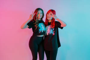 兩位擁有彩色頭髮的女性在霓虹燈工作室中微笑並做出和平手勢。