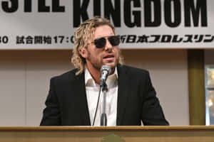 身穿西裝、戴著太陽眼鏡的男子在帶有「KINGDOM」橫幅的記者會上發言。