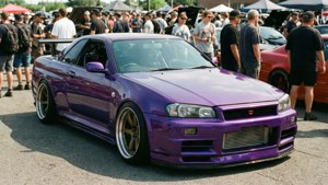 白天車展中，一輛紫色日產Skyline R34雙門轎跑車配備金色輪圈，周圍有人群。