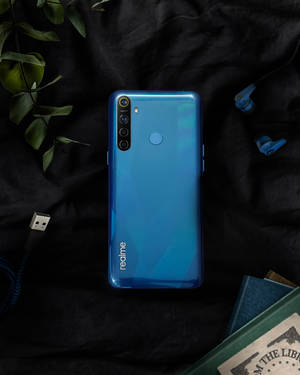 黑色布料上帶有耳塞和書本的藍色 realme 手機。 — Realme 壁紙