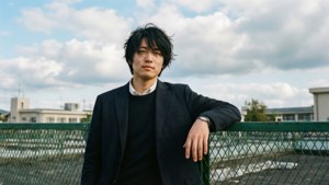 年輕男子身穿深色西裝在戶外陰天下靠著綠色圍欄
