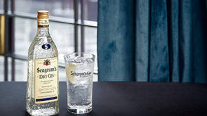 Seagram's氈酒瓶和加冰和檸檬的玻璃杯放在窗戶和藍色窗簾附近的深色桌子上。