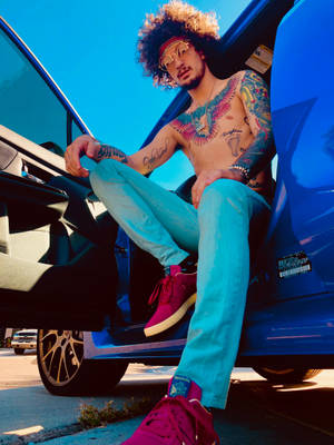 戴太陽眼鏡的紋身男子靠在一輛藍色汽車旁，穿著藍綠色褲子和粉紅色運動鞋。