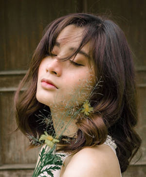 一位閉上眼睛的女子，在木牆前輕輕將野花靠近臉龐。