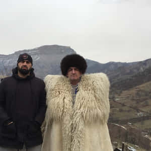兩個穿著冬季服裝的男人在白雪覆蓋的山脈前擺姿勢，其中一個穿著傳統的毛皮大衣和帽子。