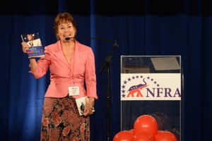穿著粉紅色西裝外套的女性在政治活動講台上拿著書，旁邊有紅色氣球。 — 粉紅 壁紙