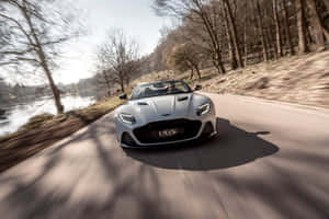 銀色Aston Martin DBS在靠近河流的道路上飛馳，周圍有光禿禿的樹木。 — Surface Laptop 壁紙