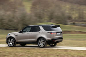 銀色 Land Rover Discovery SUV 在綠色山丘間的鄉村道路上行駛。 — MacBook Pro M2 壁紙