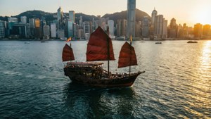 維多利亞港上一葉槳帆船悠然漂浮，城市天際線倒影與金色夕陽交相輝映的空中俯瞰景觀。
