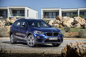 藍色BMW X1 SUV停泊在現代房屋前，有沙漠岩石和晴朗天空。 — MacBook Pro 16 壁紙