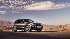 深灰色BMW X5 SUV停泊在沙漠公路上，背景有山脈和雲朵。