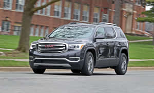 深灰色 GMC Acadia SUV 在磚砌建築和樹木附近的城市街道上行駛。 — MacBook Pro M1 壁紙