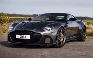 啞光黑色 Aston Martin DBS1 跑車停泊在鄉村道路上，有草地山丘。 — Steam Deck 壁紙