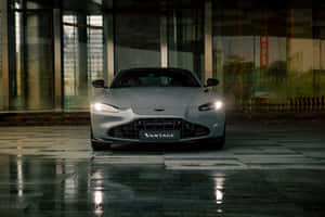 銀色Aston Martin Vantage夜間停在濕漉漉的路面上，映照出玻璃建築的倒影。 — Surface Pro 壁紙