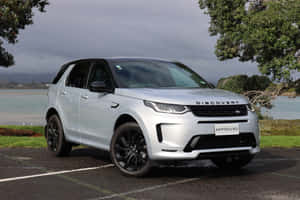 銀色 Land Rover Discovery SUV 在多雲的天空下停泊在水邊。 — MacBook Air M3 壁紙