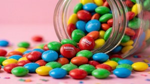 一個翻倒的玻璃罐將彩色Nestle Smarties糖果灑在粉紅色表面上，伴隨著五彩紙屑。