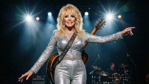 Dolly Parton 身穿銀色連身衣在舞台上表演，手持結他。