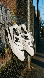 白色 New Balance 運動鞋掛在鐵絲網圍欄上，背景有塗鴉。