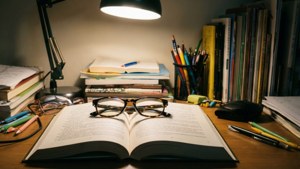 打開的書上放著眼鏡，周圍有書本、鉛筆和一盞燈在木桌上