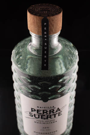 一個帶有木蓋和黑色標籤的紋理綠色Raicilla Perra Suerte瓶。
