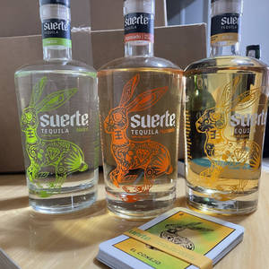 三瓶帶有兔子藝術的Suerte龍舌蘭酒瓶和一副品牌卡牌放在木質表面上。
