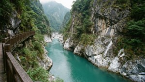 一條木徑沿著碧綠色的河流穿過陡峭的岩石峽谷，兩旁綠樹成蔭。