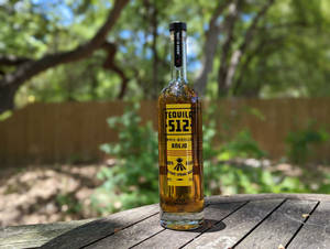 一瓶Tequila 512 Añejo放在木桌上，陽光普照的戶外環境，有樹木。