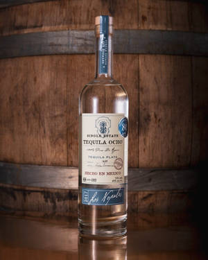 一瓶標有 Single Estate Tequila Ocho 的透明龍舌蘭酒瓶放在木桶前的木質表面上。