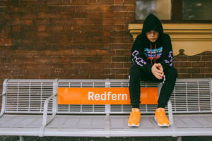 一個穿著黑色連帽衫和橙色運動鞋的年輕人坐在Redfern車站的長椅上。