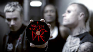 手拿著一張黑色CD，上面有紅色文字「Invaders Must Die」，前面是模糊的樂隊。