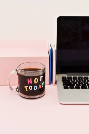 一個咖啡杯帶有「NOT TODAY」文字，旁邊有手提電腦和書本，放在粉紅色桌子上。