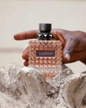 一隻手拿著 Valentino 香水瓶，瓶身帶有紋理玻璃，內有玫瑰金色液體。