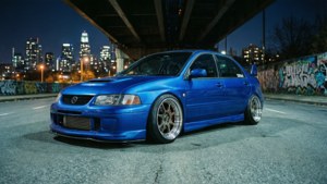 藍色改裝轎車夜間停泊在城市高架橋下，帶有塗鴉牆和天際線。