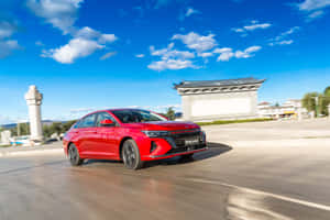 紅色轎車在晴朗天空下靠近傳統建築的道路上行駛。 — MacBook Air 15 壁紙