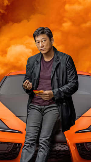 穿黑色外套的男子坐在橙色跑車引擎蓋上，橙色天空