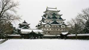 一座歷史悠久的日本城堡被白雪覆蓋，有光禿禿的樹木和遊客在附近行走。