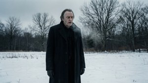 一位身穿黑色外套的長者站在雪地上，周圍有光禿禿的樹木和灰濛濛的天空。