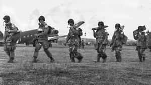 六名微笑的士兵全副武裝在田野上行軍，身後有一架飛機。