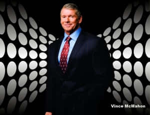 Vince McMahon 身穿深色西裝和紅色領帶，站在帶有白色橢圓燈光的黑色牆壁前。