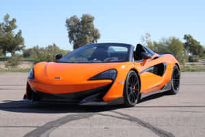 橙色McLaren 570S Spider開篷車停泊在瀝青路上，背景有樹木和藍天。 — MacBook Pro 14 壁紙